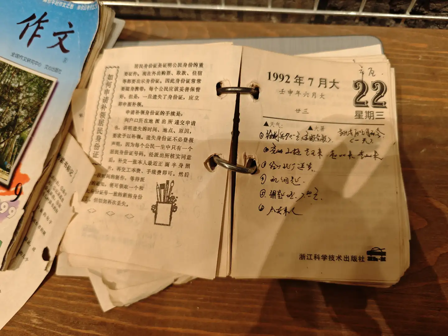 一本打开的日历显示1992年7月22日，旁边有一本作文书。日历上记录了当天的天气和几项活动。页面设计简洁，字体清晰。