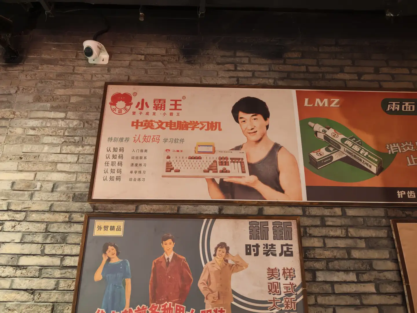 墙上挂着两幅广告海报，上方海报展示“小霸王 中英文电脑学习机”，下方海报宣传时装店。海报色彩鲜艳，人物形象生动。背景为砖墙，上方有监控摄像头。