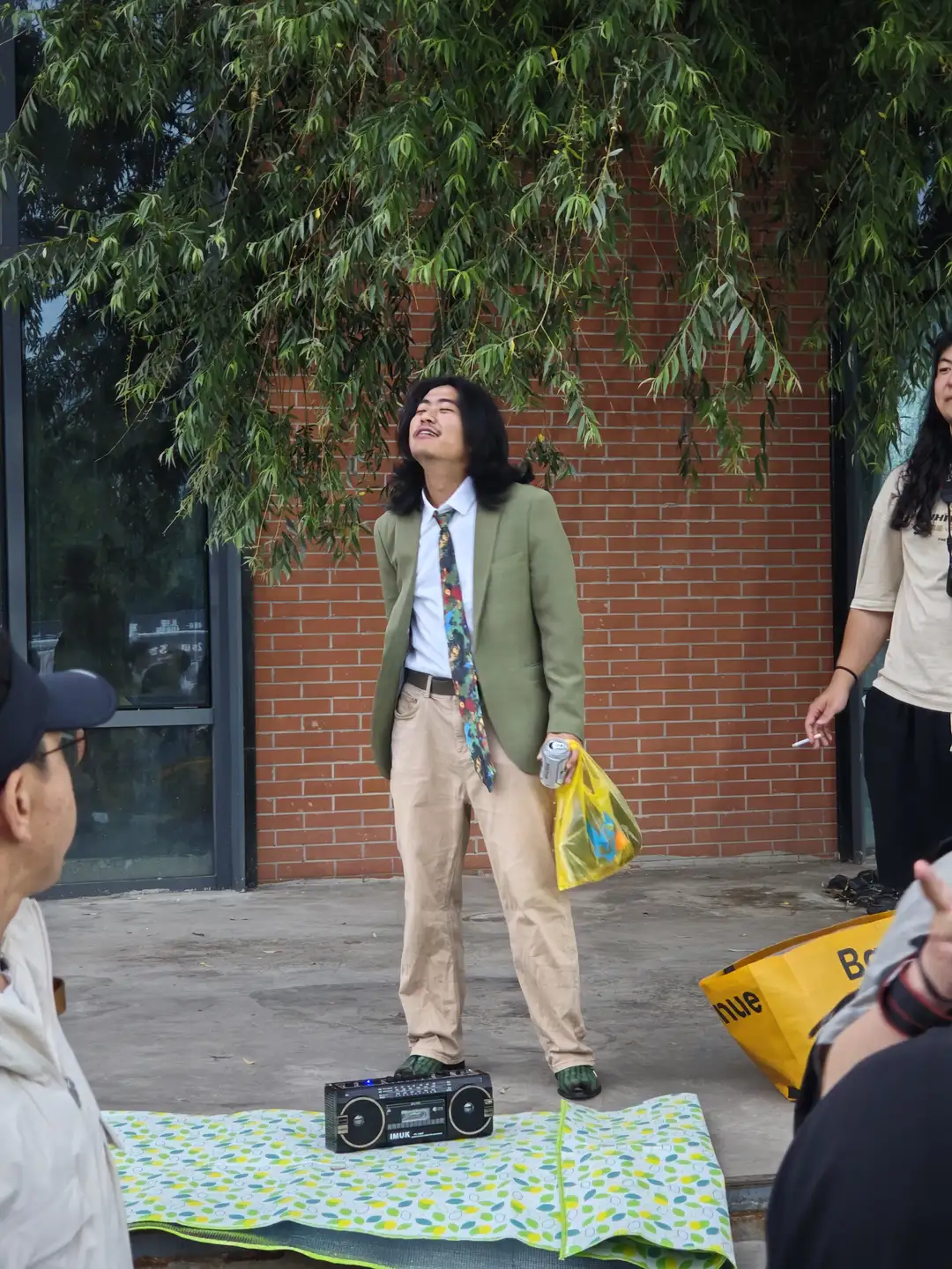 一名男子站在户外，手持塑料袋和罐装饮料，旁边放置一台老式收音机。男子穿着绿色西装外套和卡其裤，背景为红砖墙和垂柳。旁边有几个人在观看。