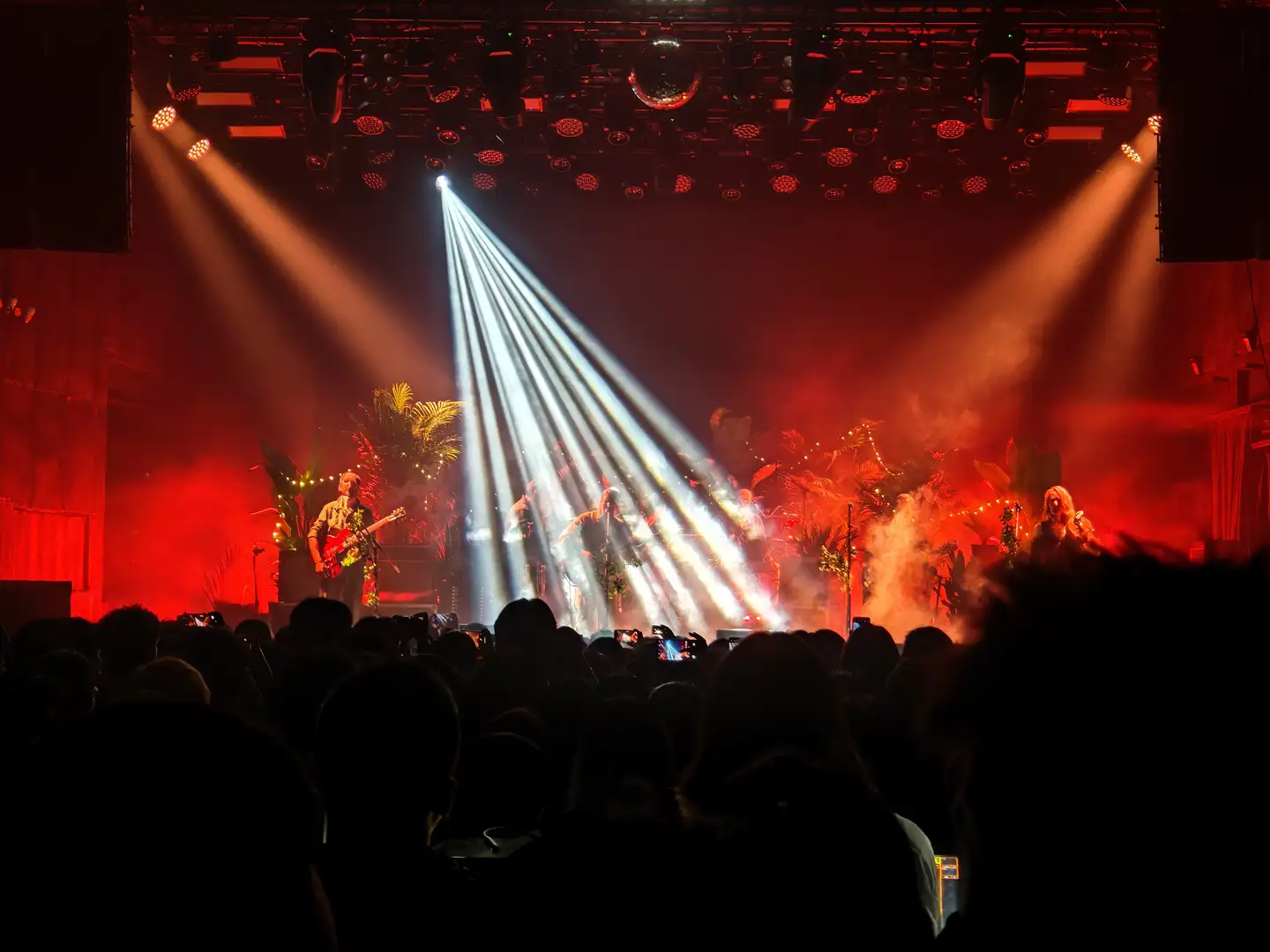 舞台上灯光璀璨，乐队正在表演，观众席人头攒动。舞台布置有热带植物，灯光效果炫目。观众用手机拍摄表演场景。