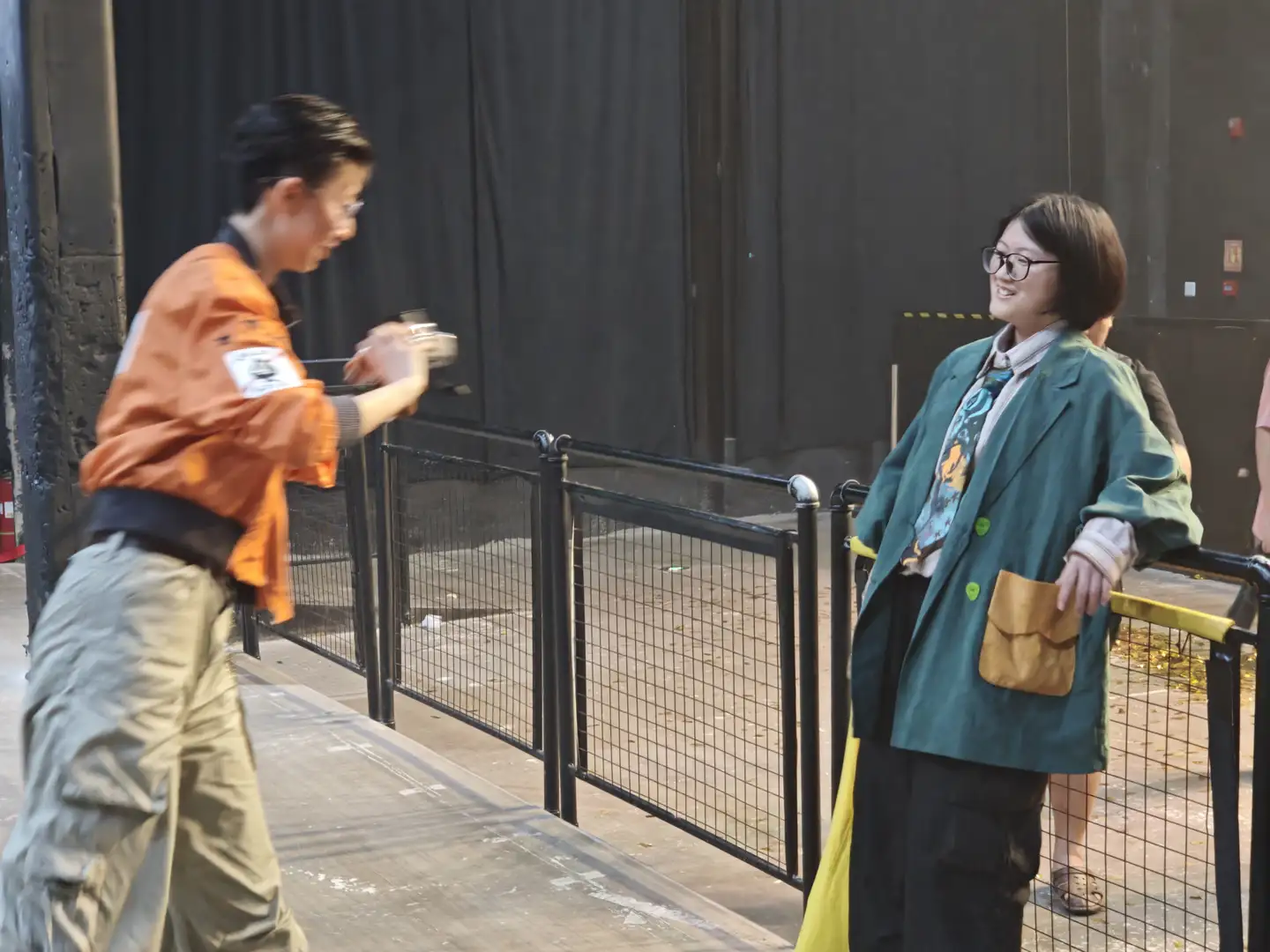 一个人穿着橙色外套正在用相机拍照，另一个人穿着绿色外套站在一旁微笑。背景是一面黑色的墙和金属围栏。场景看起来像是在户外活动区域。