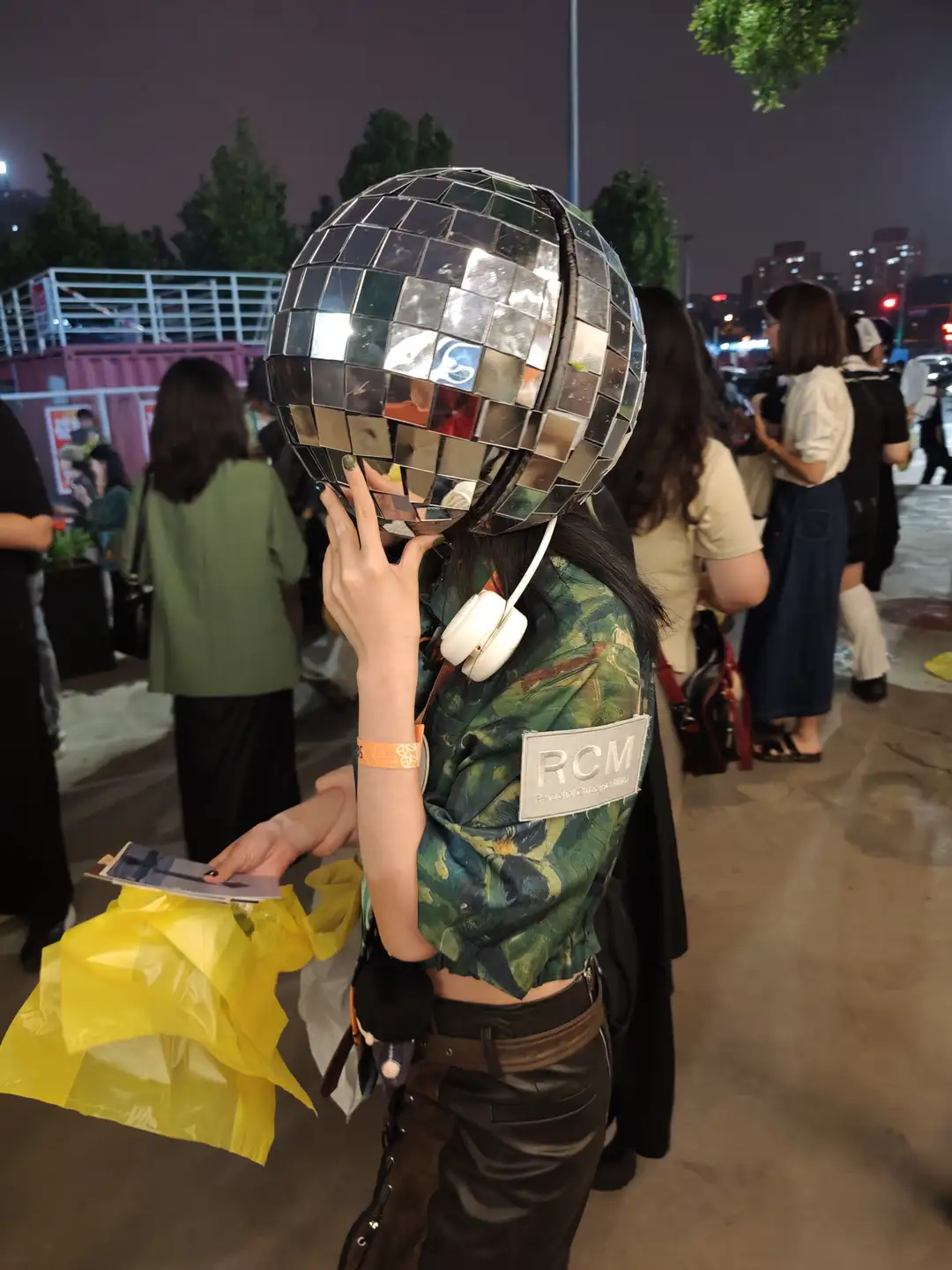 一个人戴着由镜子组成的球形头盔，穿着迷彩上衣和黑色裤子，手里拿着黄色塑料袋。背景中有多个人在行走，场景似乎是在夜晚的户外。