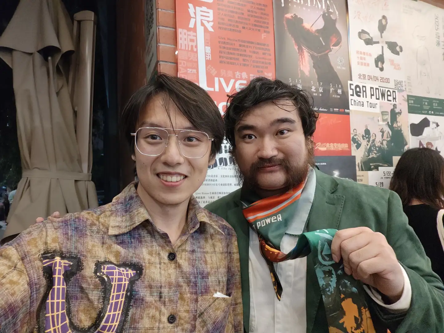 两个人站在一起合影，左边的人穿着带有紫色图案的格子衬衫，右边的人穿着绿色西装外套并系着彩色领带。背景墙上贴有多张海报，其中包括“SEA POWER China Tour”的信息。