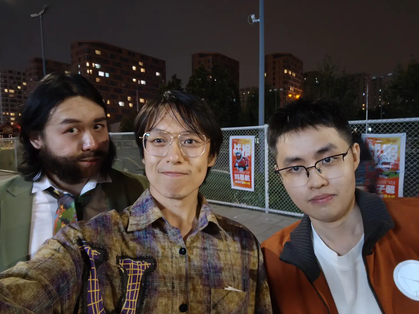 三个人站在一起自拍，左边的人穿着格子衬衫，中间的人穿着相同风格的衬衫，右边的人穿着橙色外套。背景是夜晚的城市景观和铁丝网围栏，围栏上有张贴的海报。