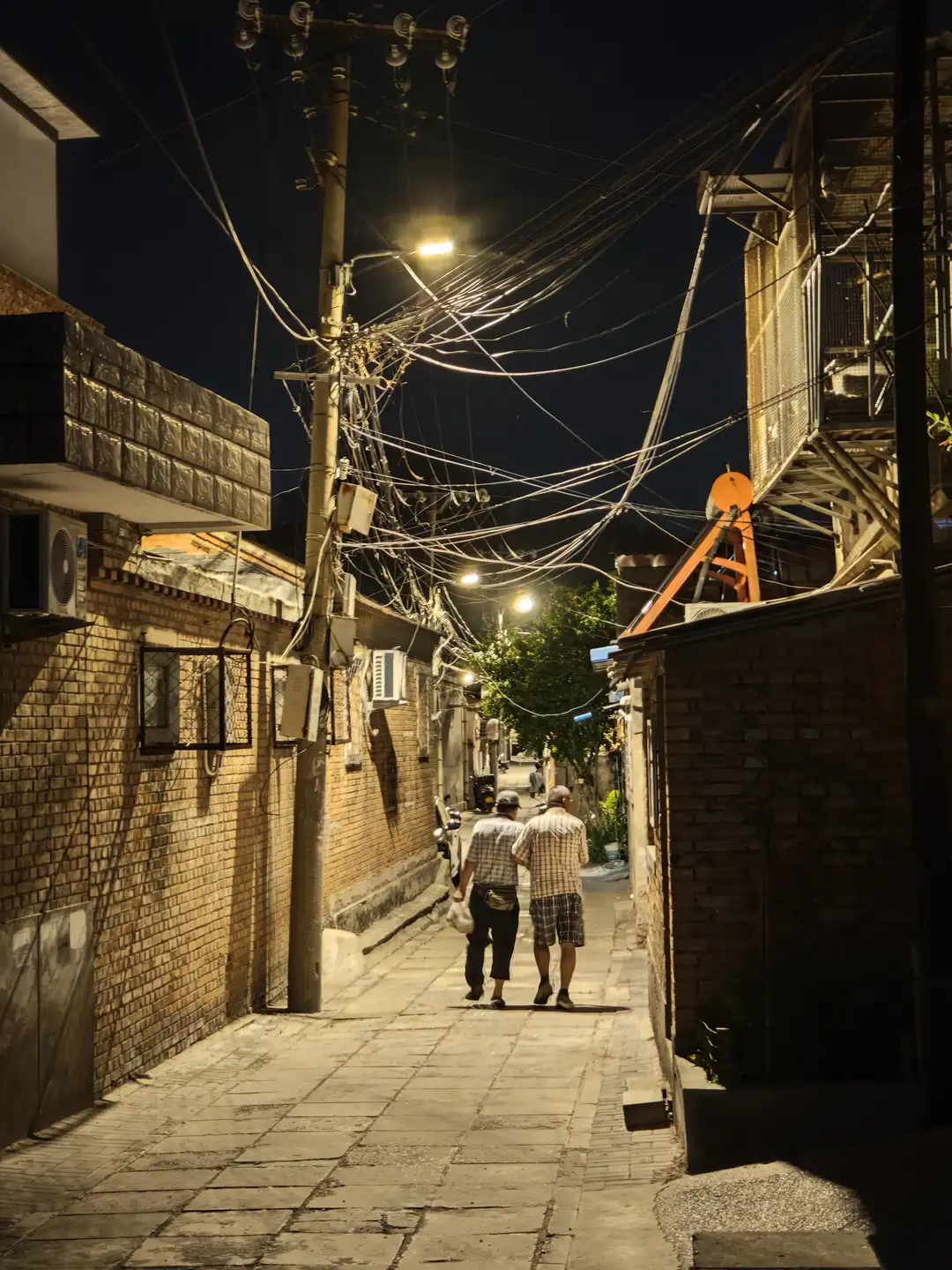 夜晚的巷子里，两个人并肩行走，周围是砖墙和电线杆。路灯照亮了他们的前方，电线在空中交错。远处有树木和更多的建筑物。