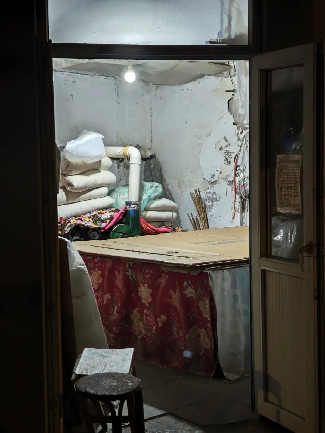 房间内部陈设简单，墙上剥落的油漆显露出岁月痕迹。门口堆放着被褥和杂物，中央有一张覆盖红色花纹布料的桌子。墙上贴着一张纸，内容为注意事项或公告。