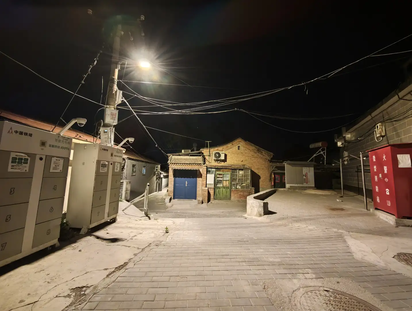 夜幕下的街道空旷，路灯明亮，照亮了前方的建筑和路面。左侧有标有“中国铁塔”字样的设备箱，右侧是一个红色消防柜。街道尽头有一栋老式建筑。