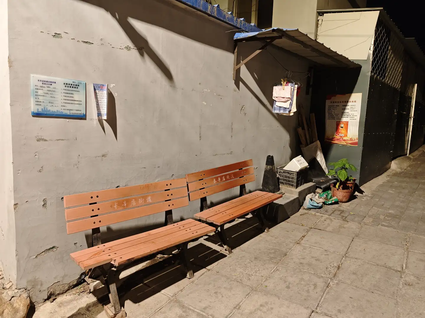 夜晚的街角，两排木质长椅靠墙放置，墙上贴有蓝色告示牌。长椅旁边有一个黑色交通锥和一些杂物。墙角处摆放着几盆绿色植物。