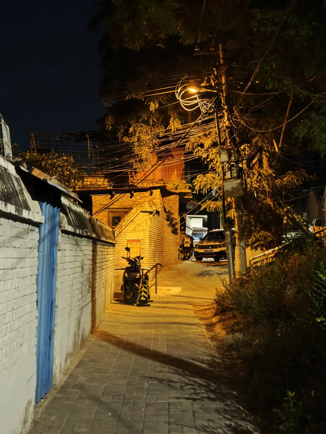 夜晚的小巷，路灯温暖的光芒洒在地面上，左侧是白色围墙和蓝色大门。右侧有树木和灌木丛，小巷尽头停放着一辆摩托车和汽车。电线在空中纵横交错。
