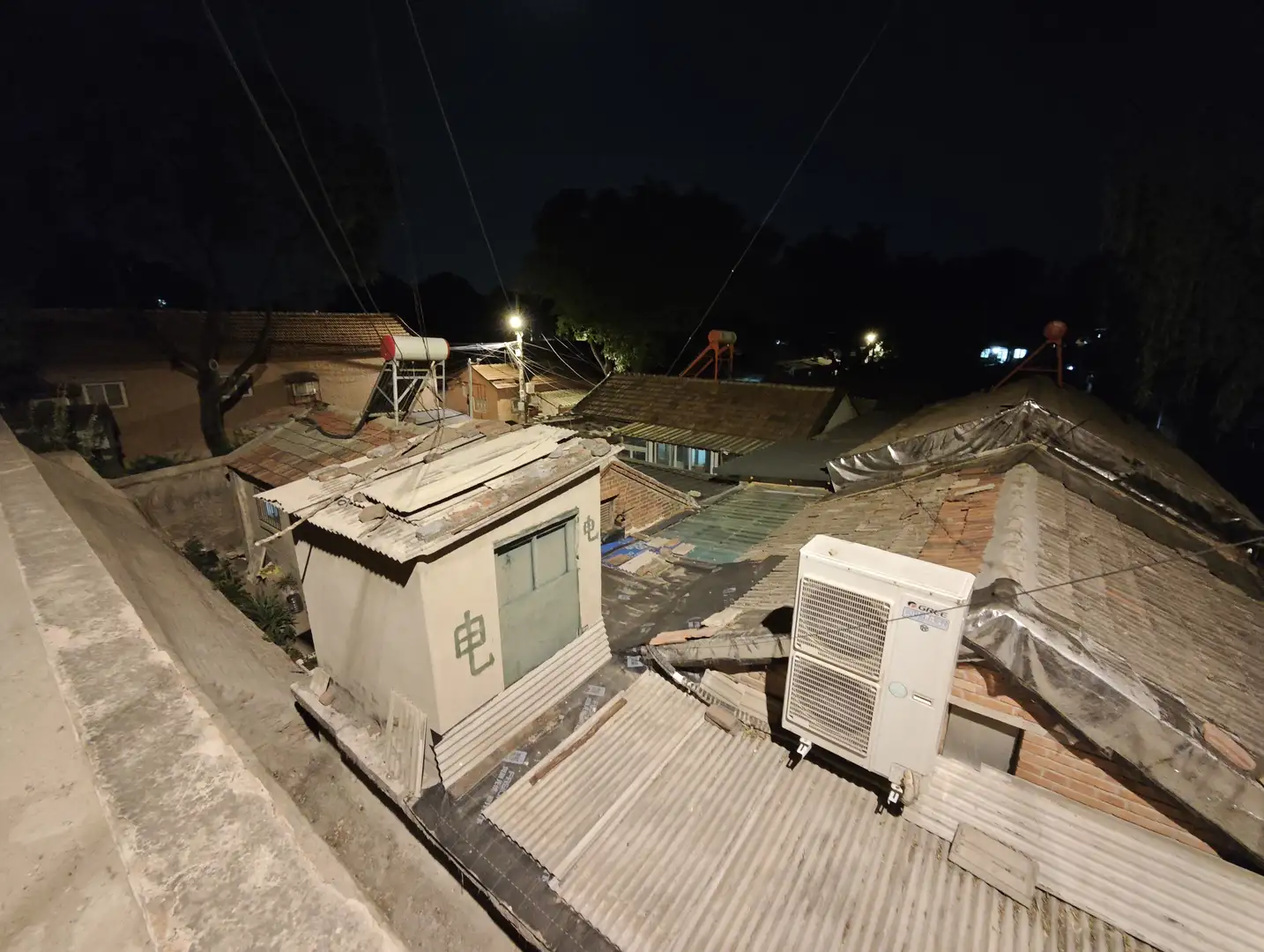 夜晚的屋顶视角，瓦片和金属板构成屋顶表面，上面放置着空调外机和储水罐。远处可以看到更多房屋和树木的轮廓。天空漆黑，几盏灯光点缀其间。