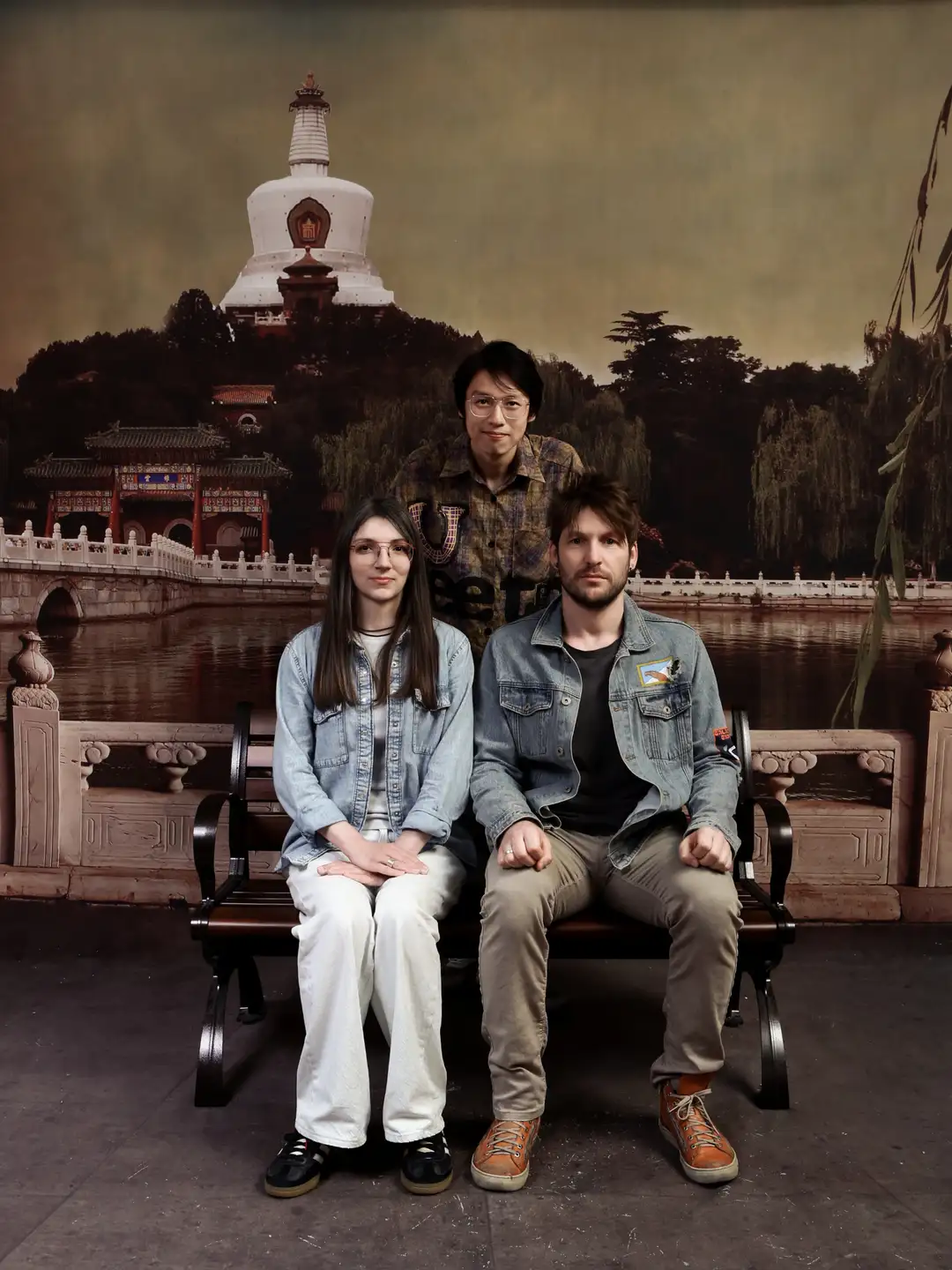 三个人坐在长椅上，前面两人坐着，后面一人站着。背景是一幅描绘传统建筑和湖泊的图画，远处有一座白色的塔楼。三人穿着休闲，姿态自然。