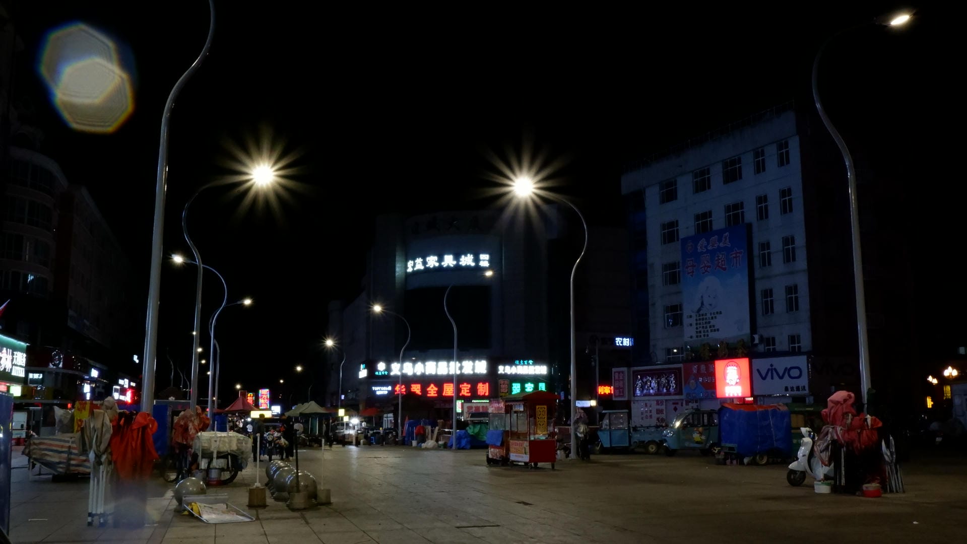 At the night market square, streetlights are on, surrounded by stalls and billboards.