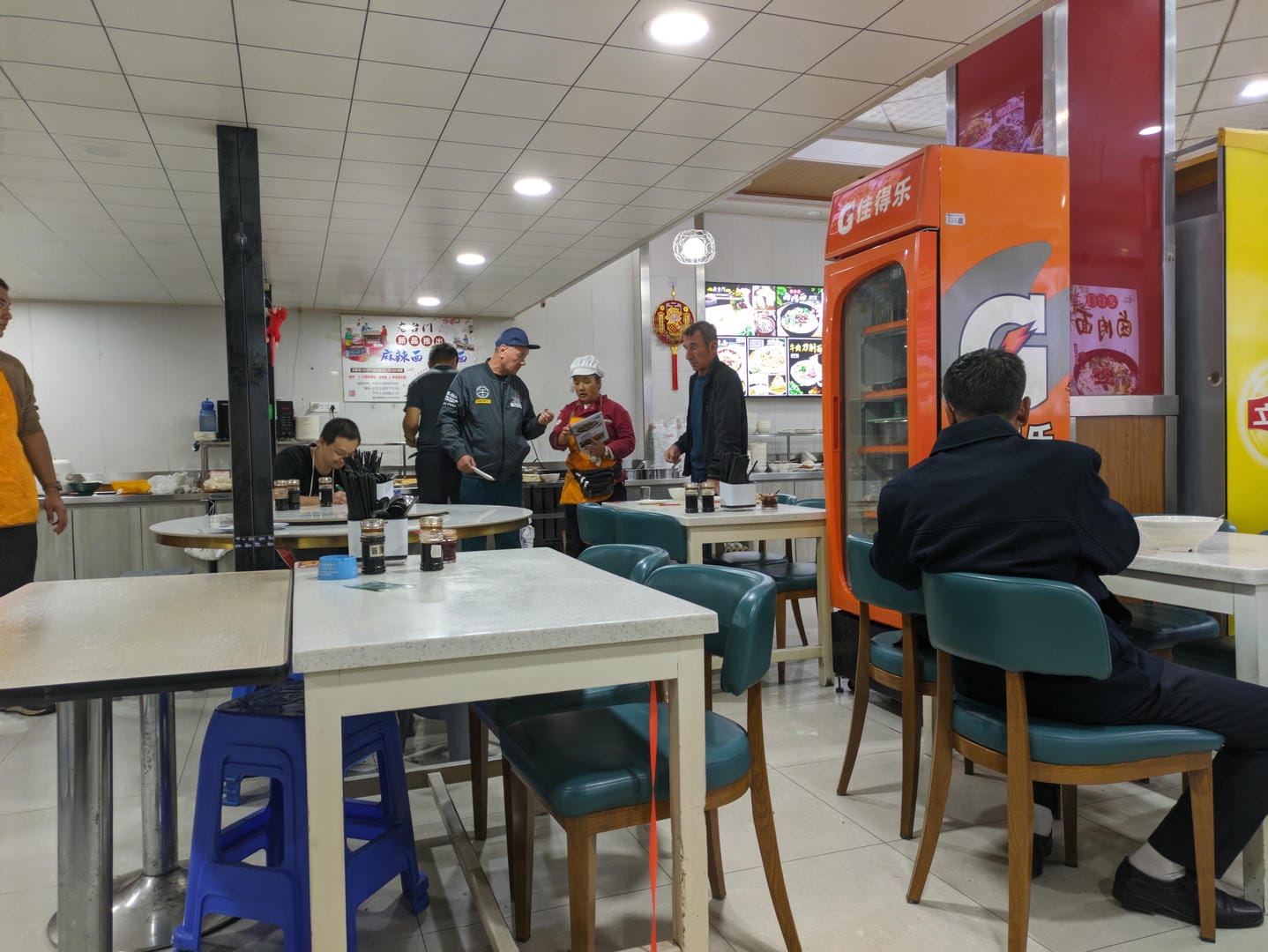 Inside a restaurant, people are dining at tables, with menus hanging on the wall.