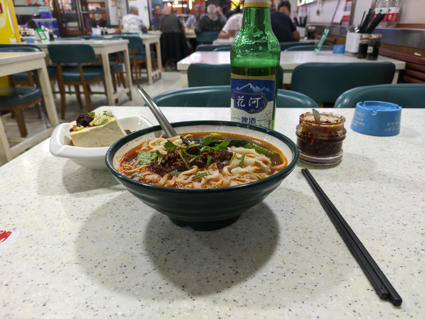 A bowl of noodles, a bottle of beer, a plate of tofu, and chili sauce on the table.