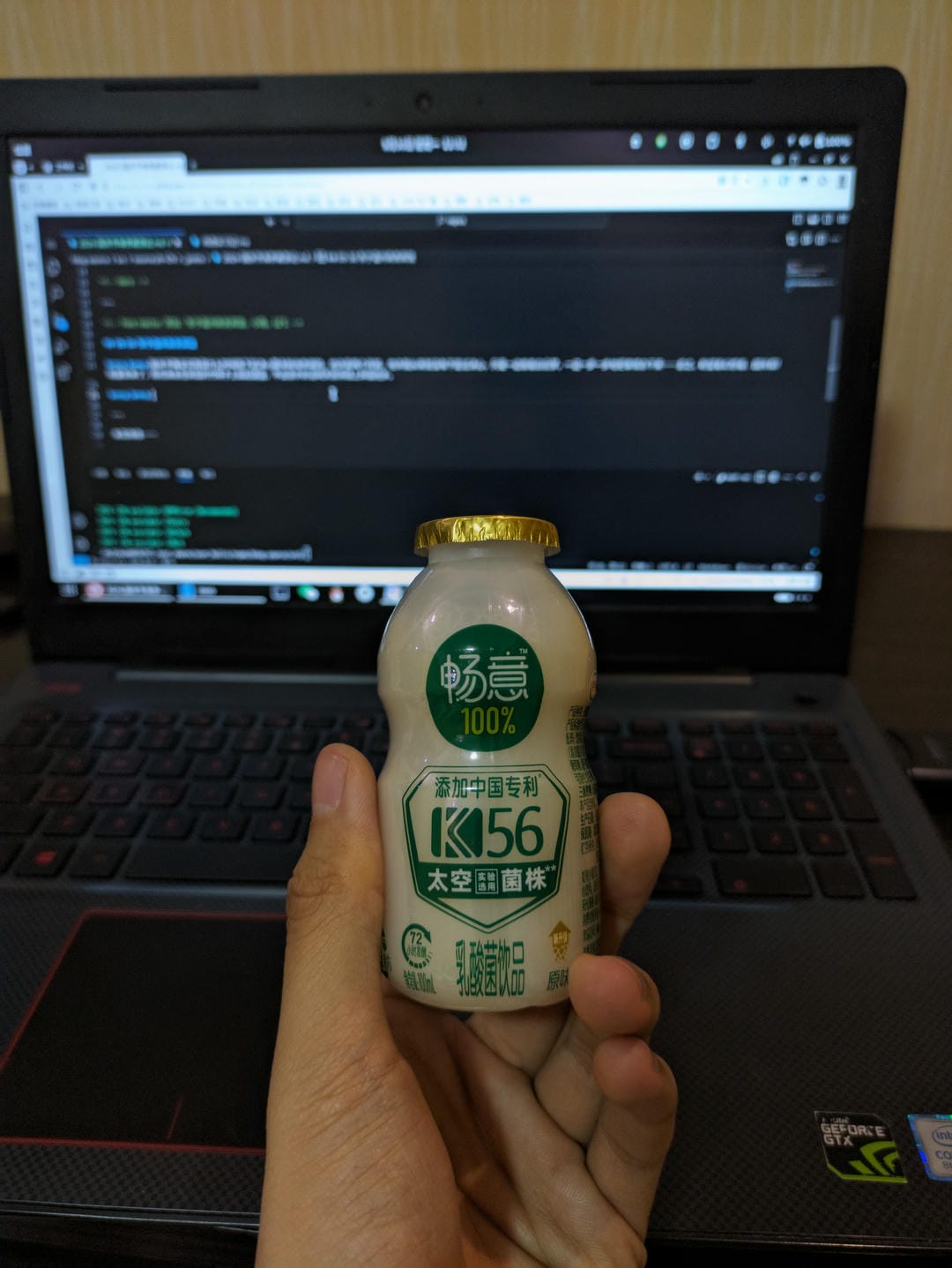 A hand holding a bottle of lactic acid bacteria drink, with a laptop in the background.