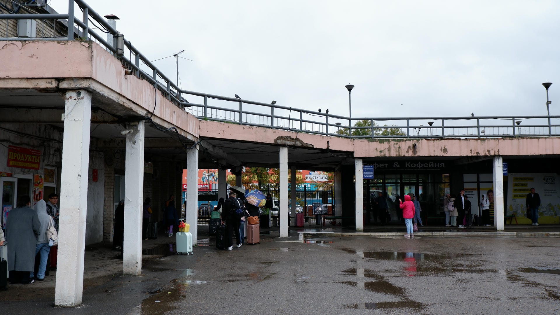 A rainy outdoor scene with people waiting in front of a building, the ground is wet.
