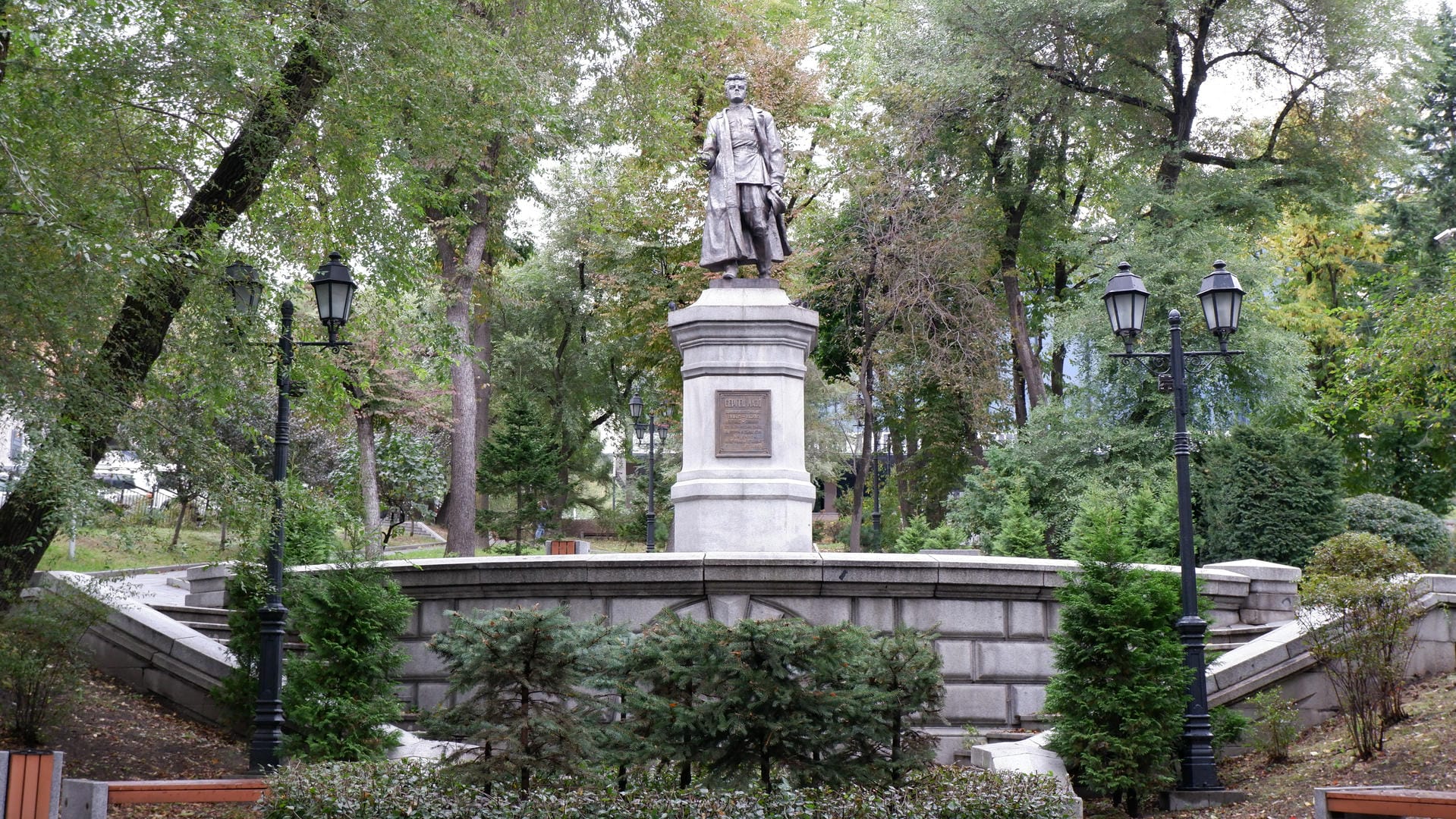 A statue at the center of a park, surrounded by trees and street lamps.