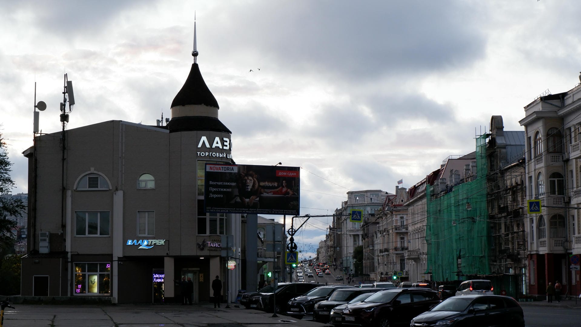 A gray building with spire-topped roof and side billboards under cloudy sky, cars lining the street.