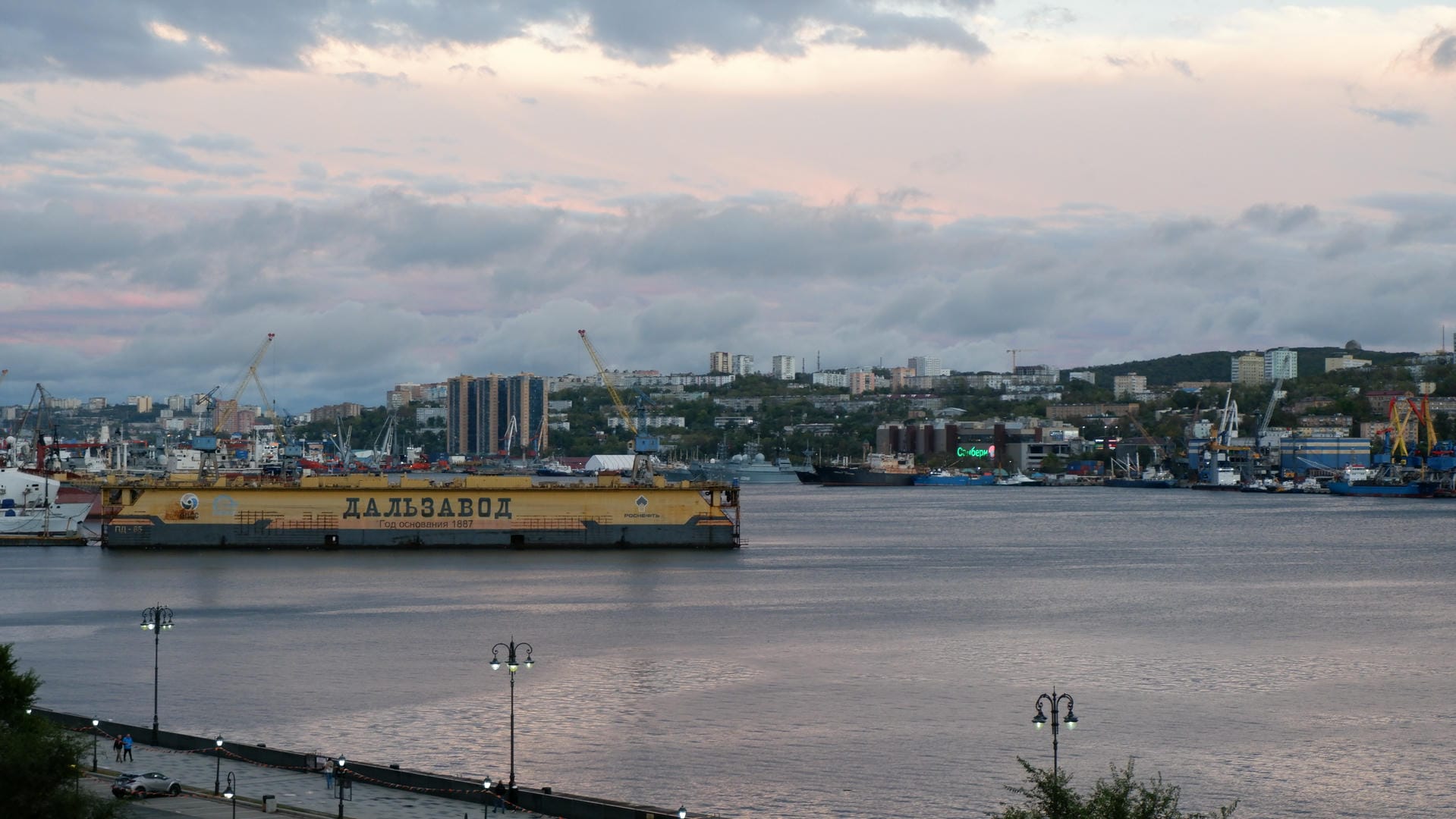 Harbor area with yellow buoy marked “ДАЛЬЗАВОД”, city buildings and cranes in background.
