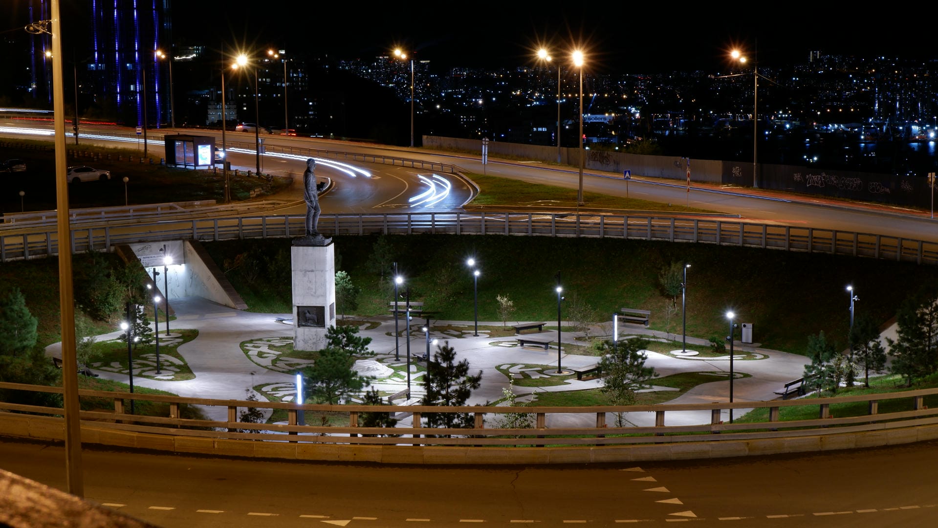 Night view of city park with central statue, illuminated paths and trees.