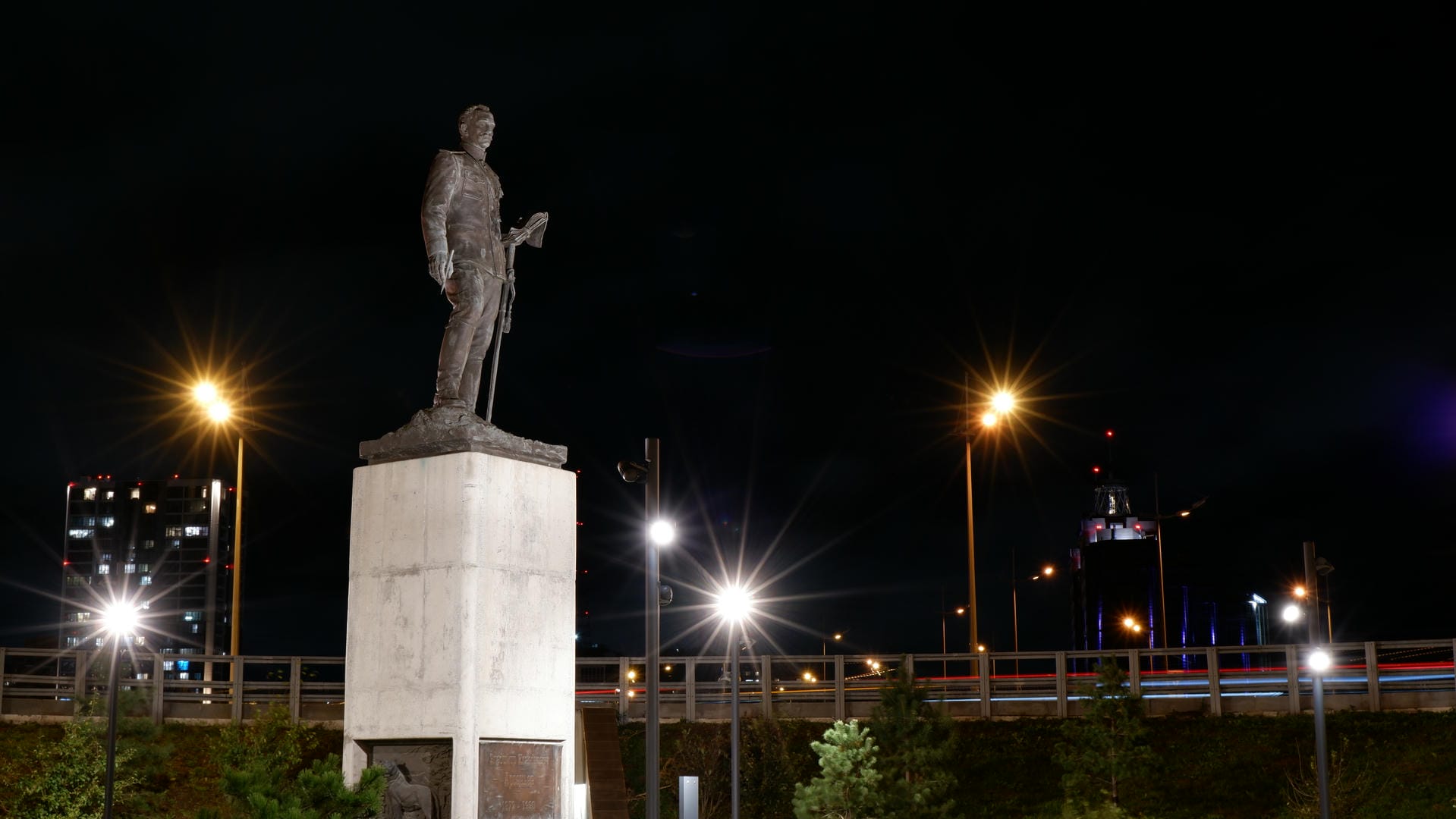 Night close-up of statue on square pedestal illuminated by street lamps.