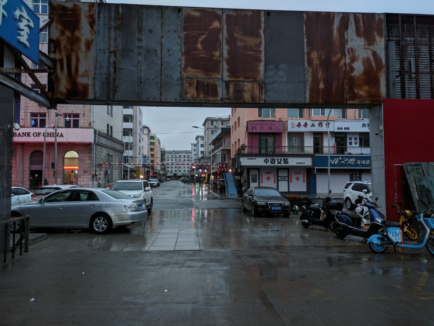 Rain-wet street with parked cars/motorcycles and overhead rusty metal structures.