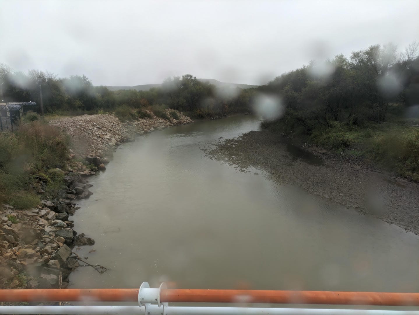 A muddy river with rocky/tree-lined banks and foreground railings.
