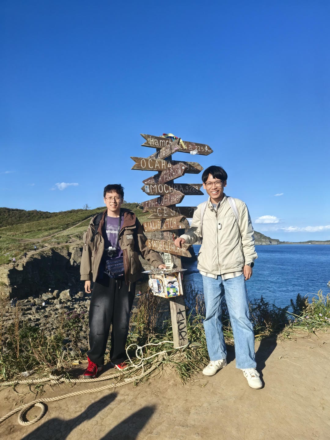 Two people standing seaside with distance sign behind. Left person wears brown jacket/black pants, right in light jacket/blue jeans.
