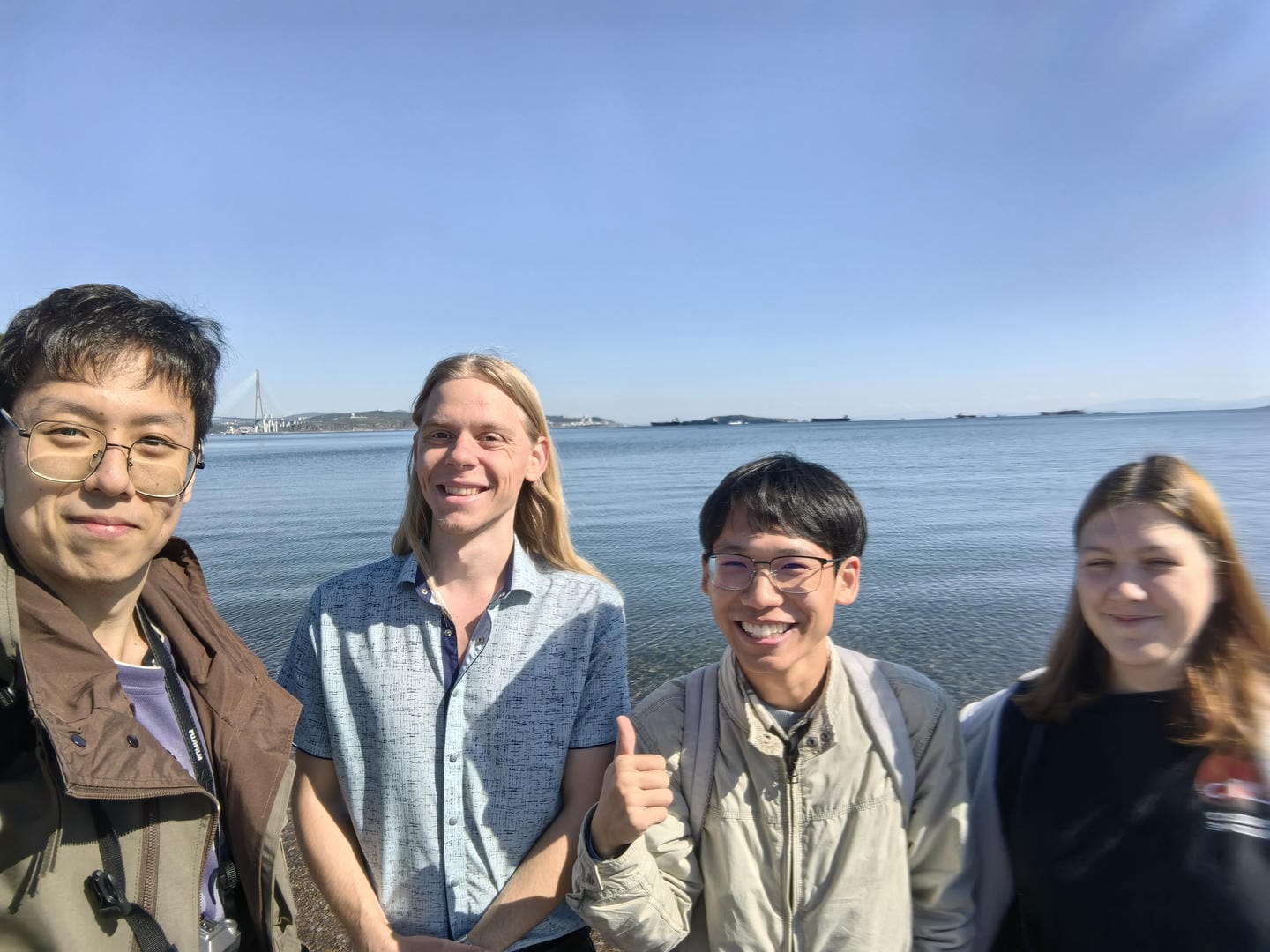 Four people by sea: bespectacled person in brown jacket; blonde in blue shirt; thumbs-up person in light jacket; brunette in dark top.