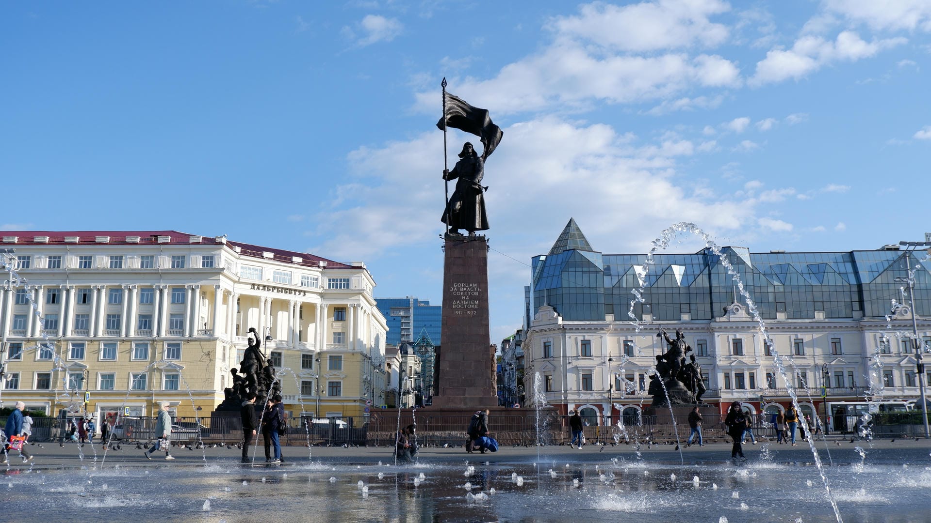 Square featuring fountain, statue holding flag, and surrounding buildings.