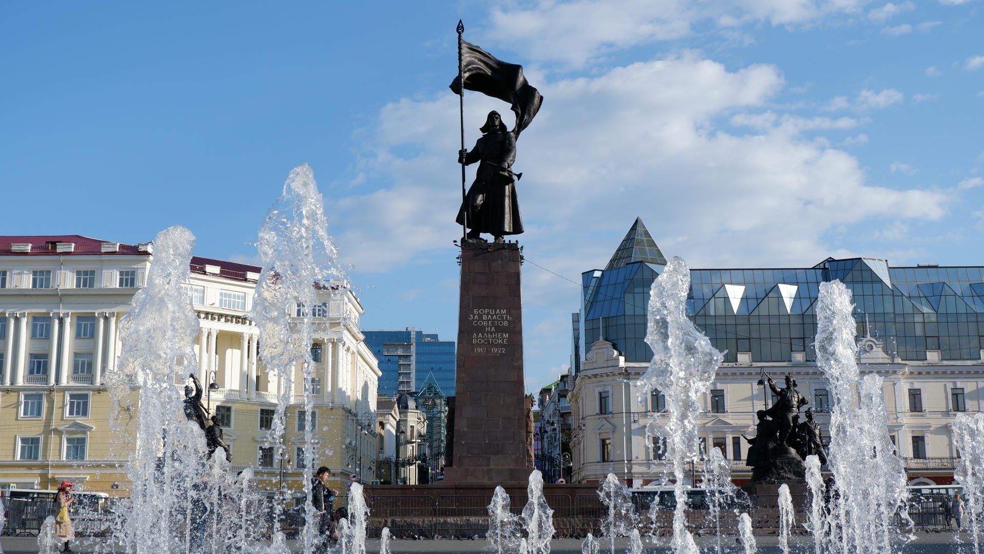 Square with fountain, flag-bearing statue and architectural structures.