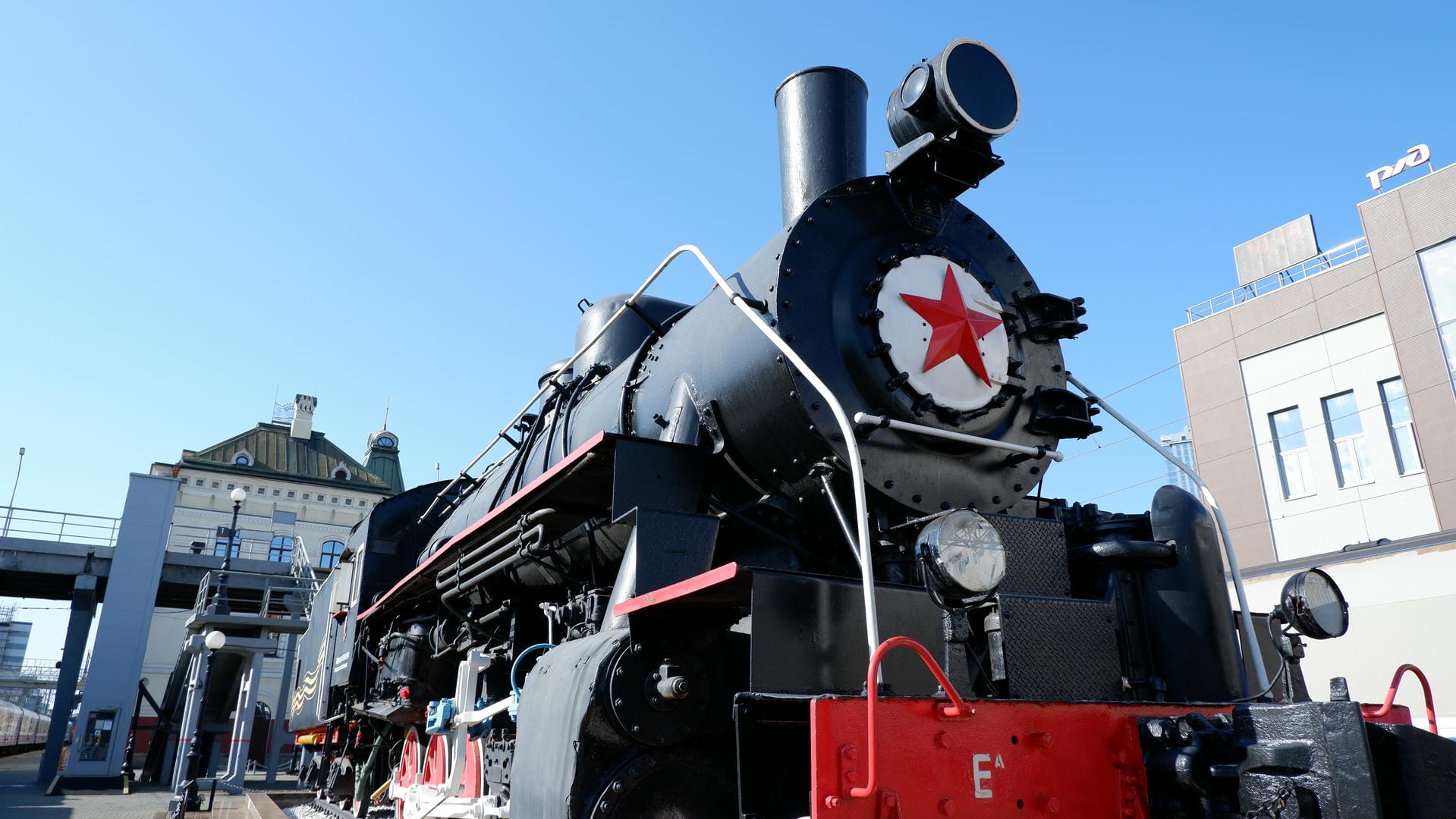 Black steam locomotive with red star emblem, modern buildings in background.