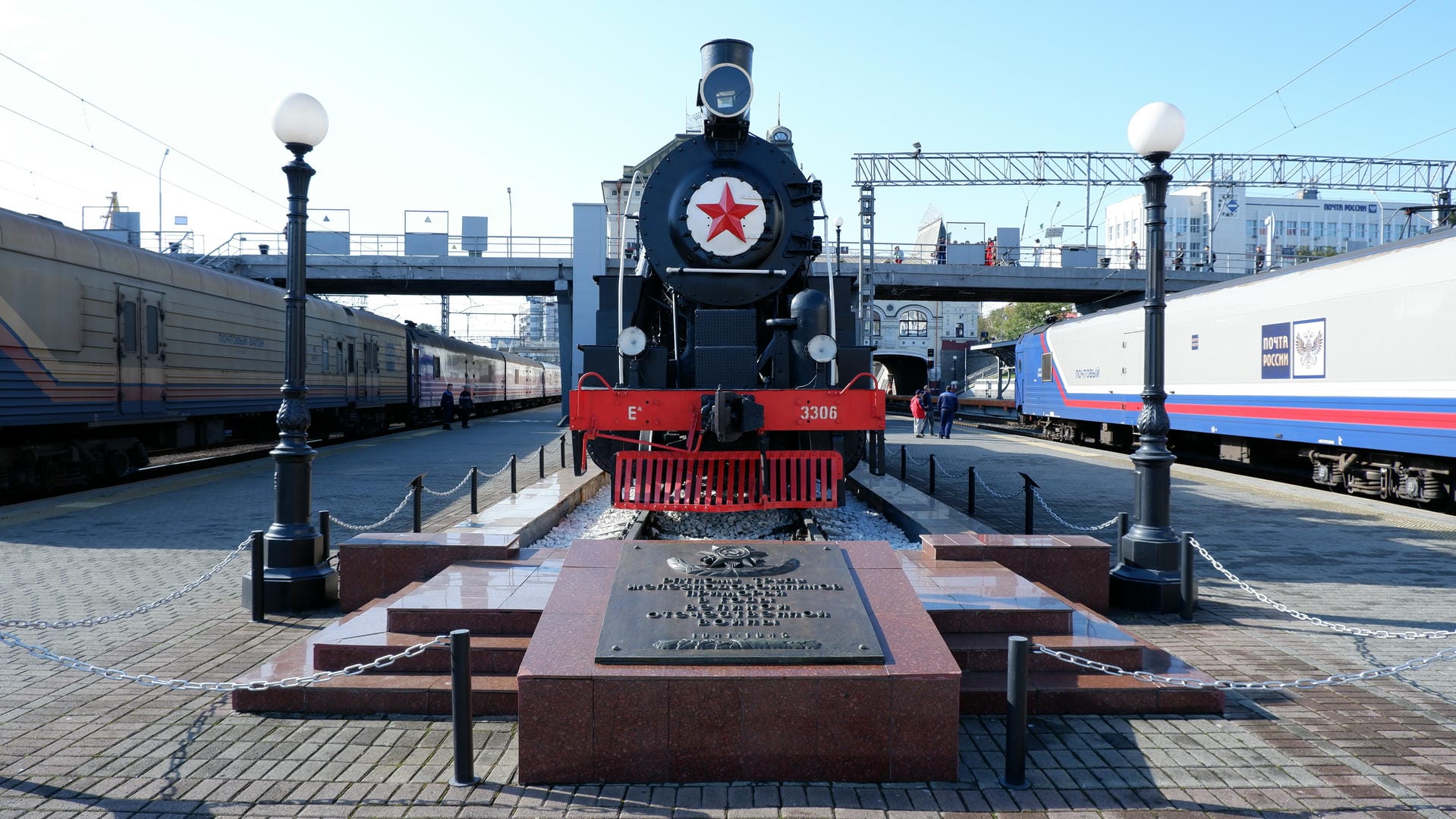 Monument placed before steam locomotive, protected by chain fence with trains parked alongside.