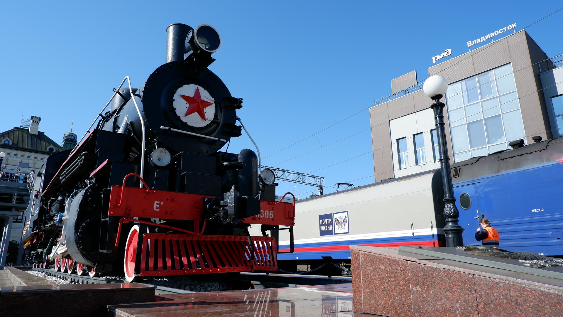 Steam locomotive displayed outdoors with red star emblem, orange-clad person standing nearby.