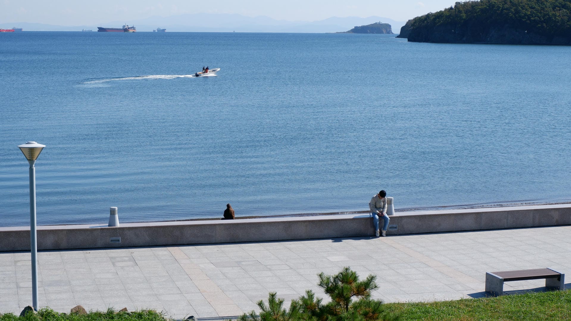 Two people sitting on seaside walkway with small boat sailing in distance.