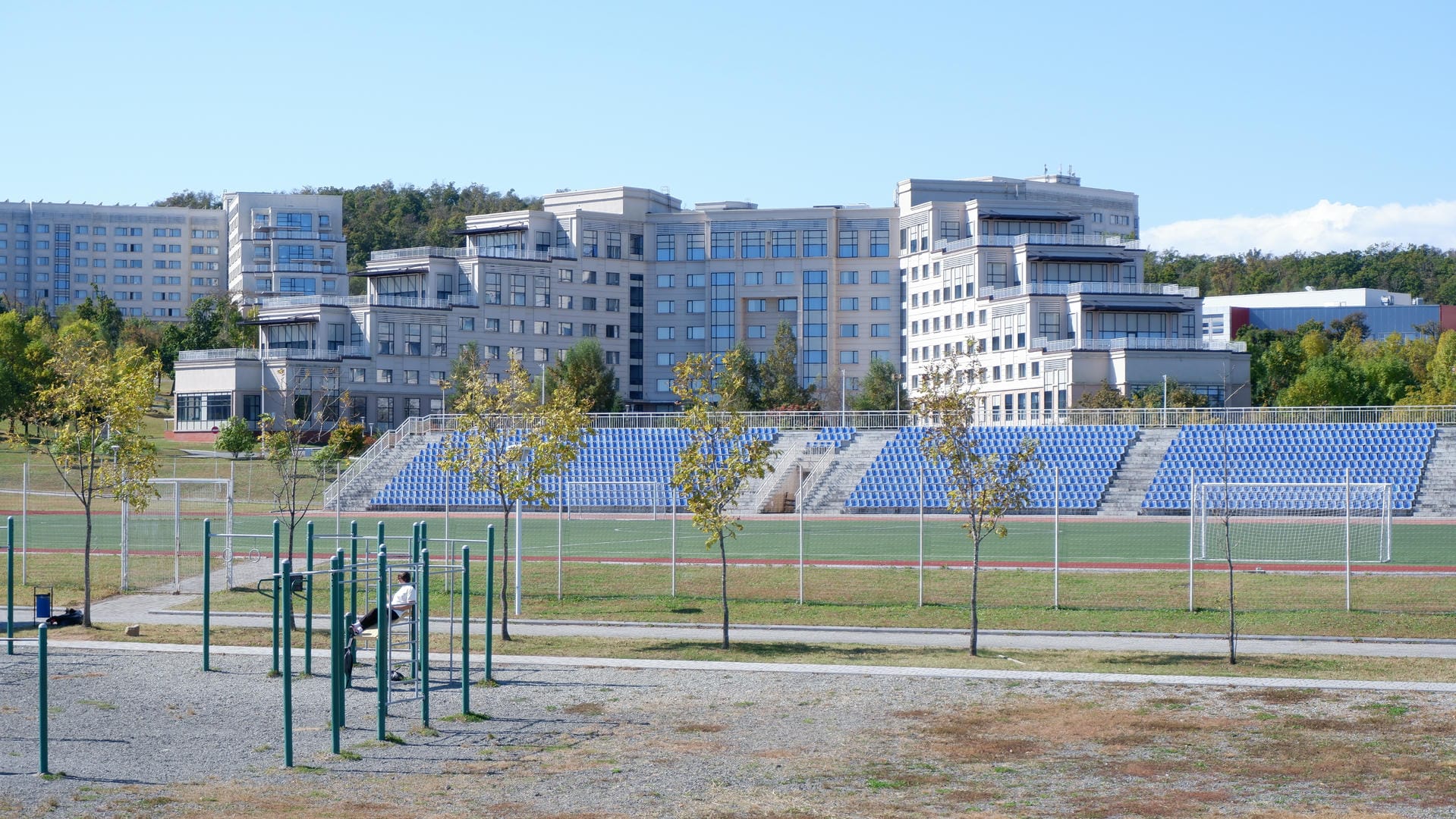 Soccer field flanked by blue seats and multi-story building in background.
