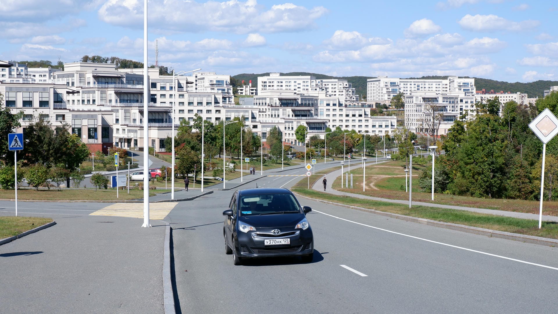 Black car driving on road with white buildings in background.