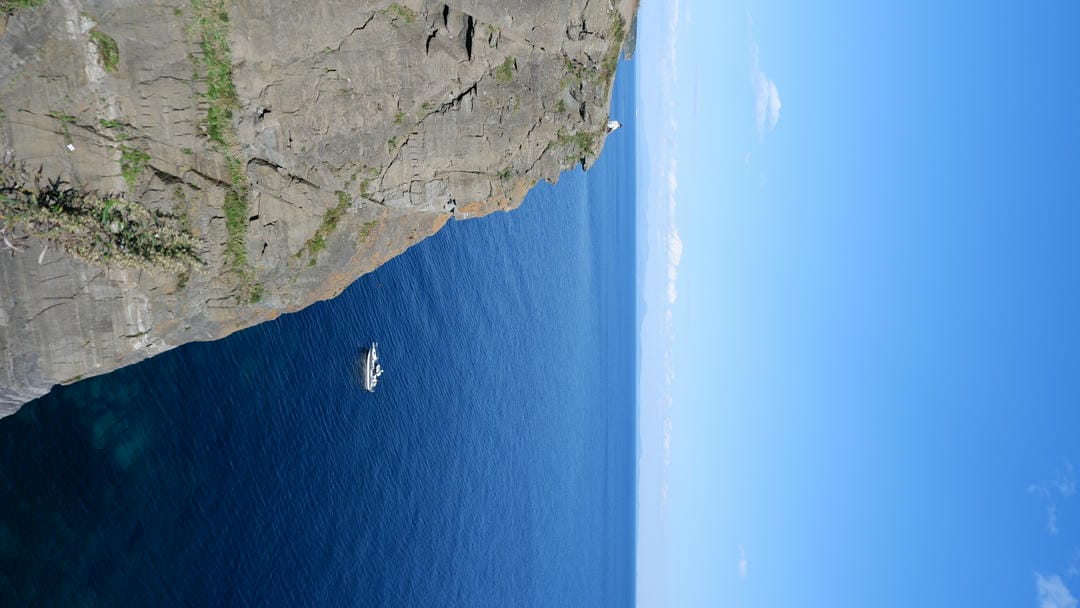 Small boat moored near cliff in deep blue sea, mountains in distance.