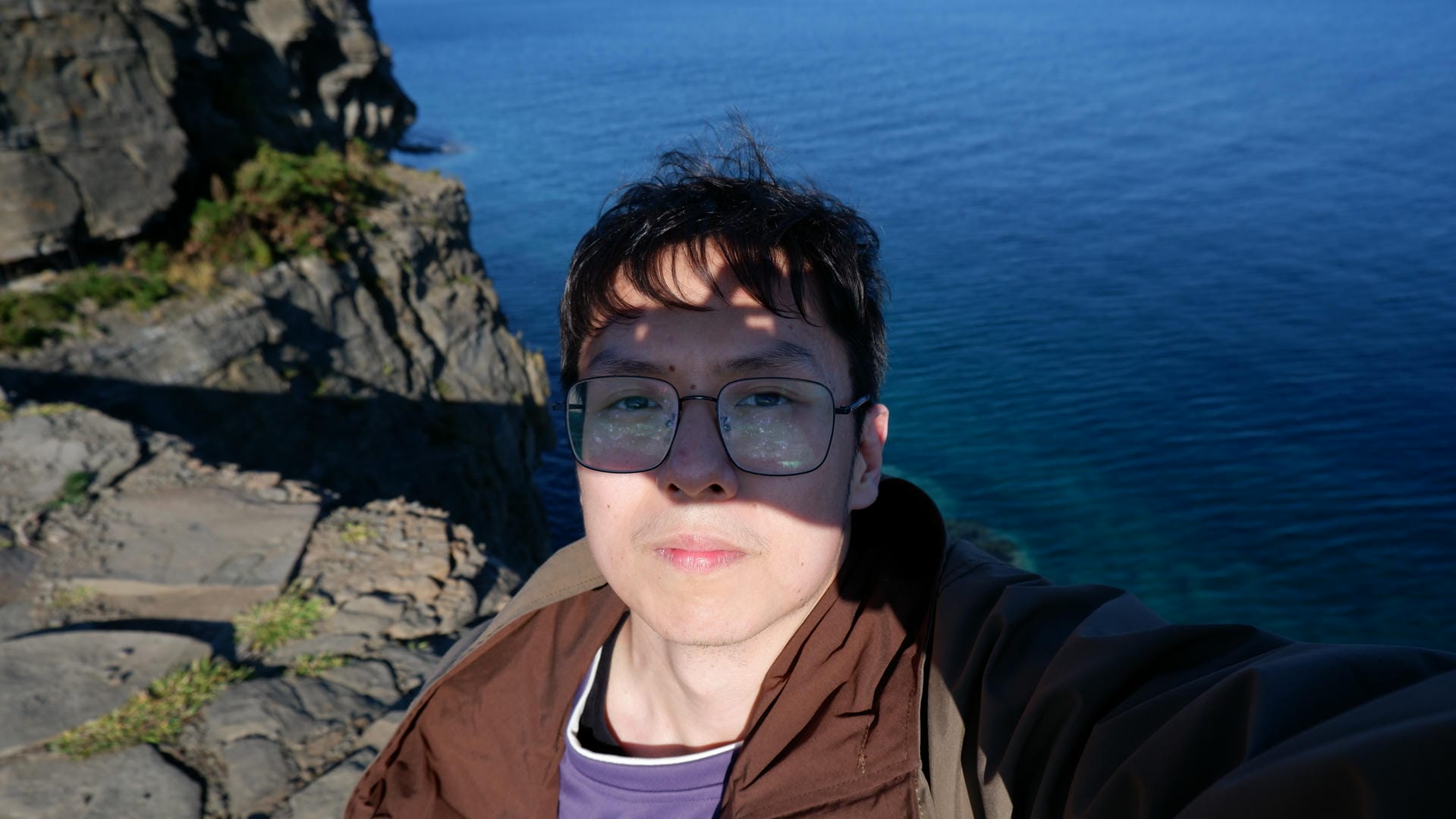 Bespectacled person standing at cliff edge with dark blue sea and rocks behind.