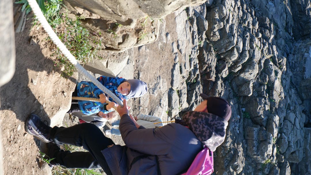 Adult assisting child in blue jacket and gray hat climbing hillside.