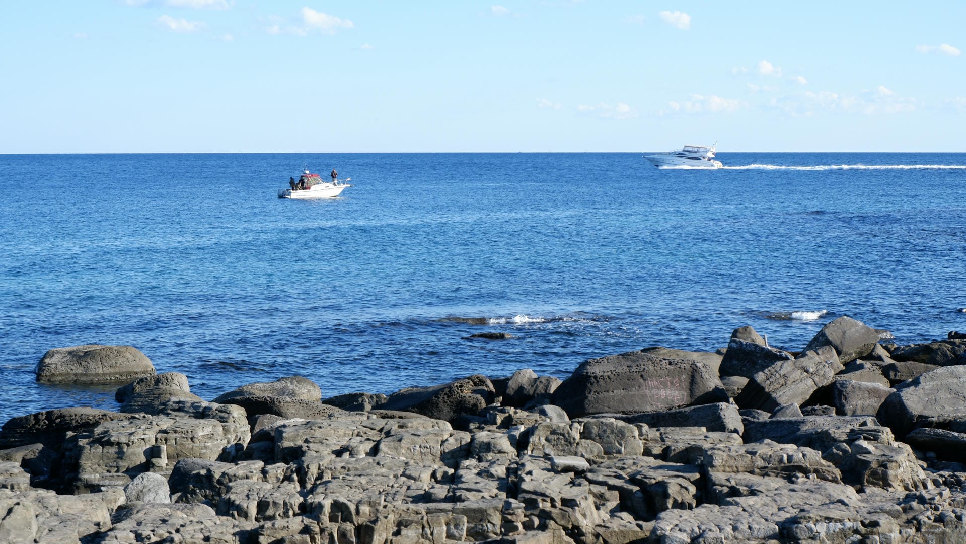 Two boats sailing on deep blue sea with rocky foreground.