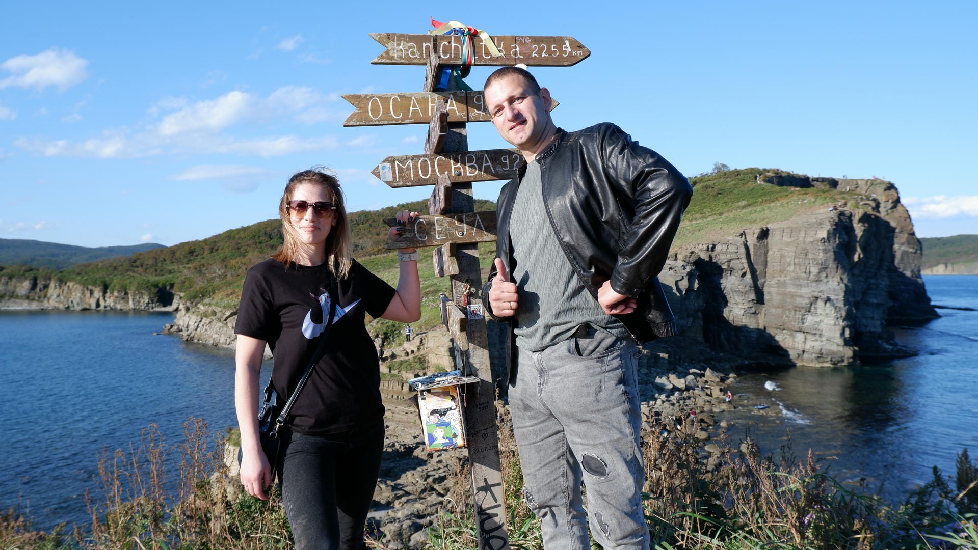 Two people standing before directional sign listing multiple cities/distances.