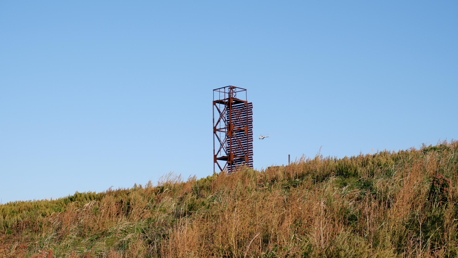 Metal tower on grassland with helicopter flying nearby.