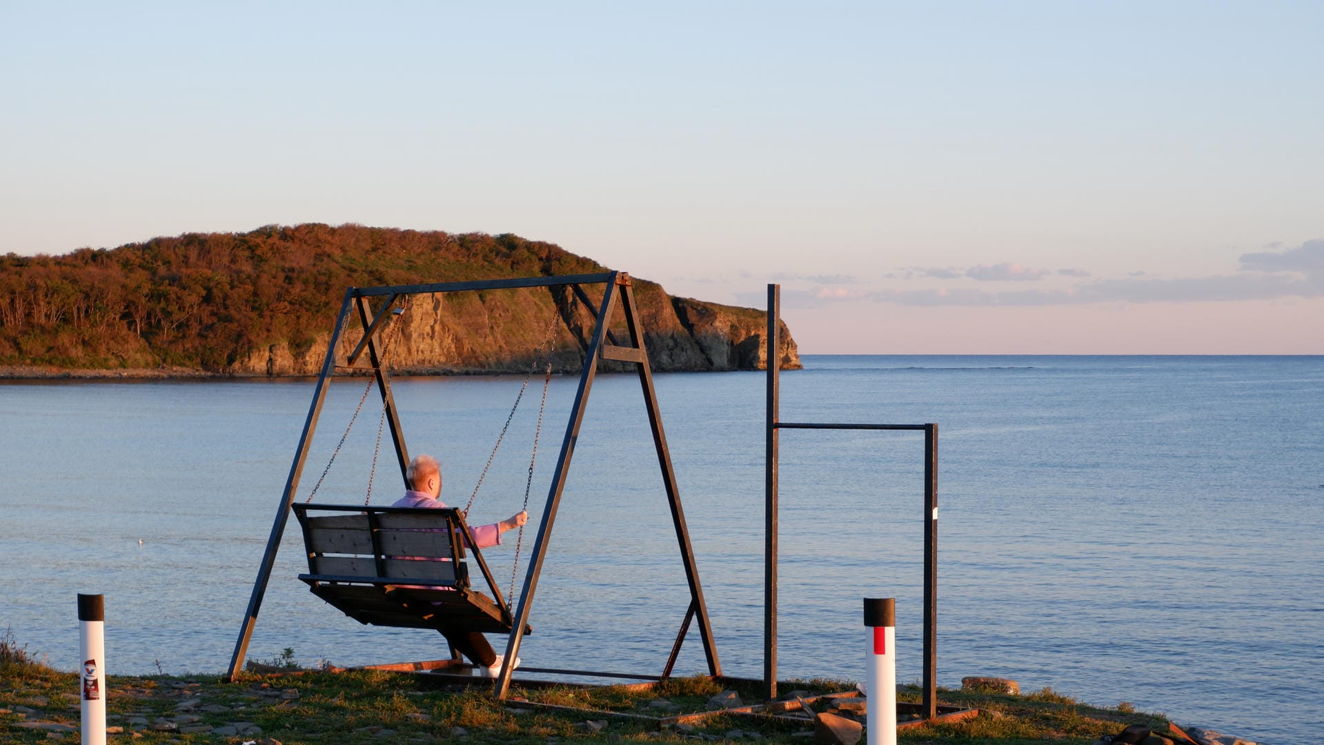 Elderly person sitting on swing facing sea with distant island.