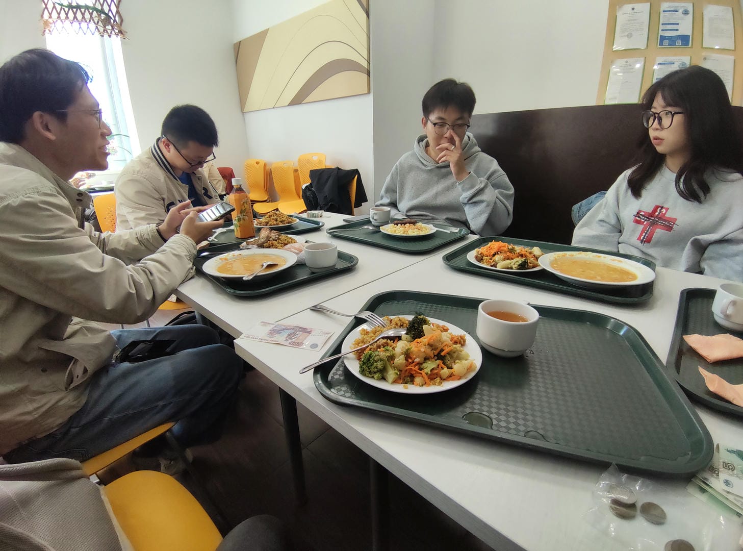 Four people at dining table with food/drinks, one using phone.