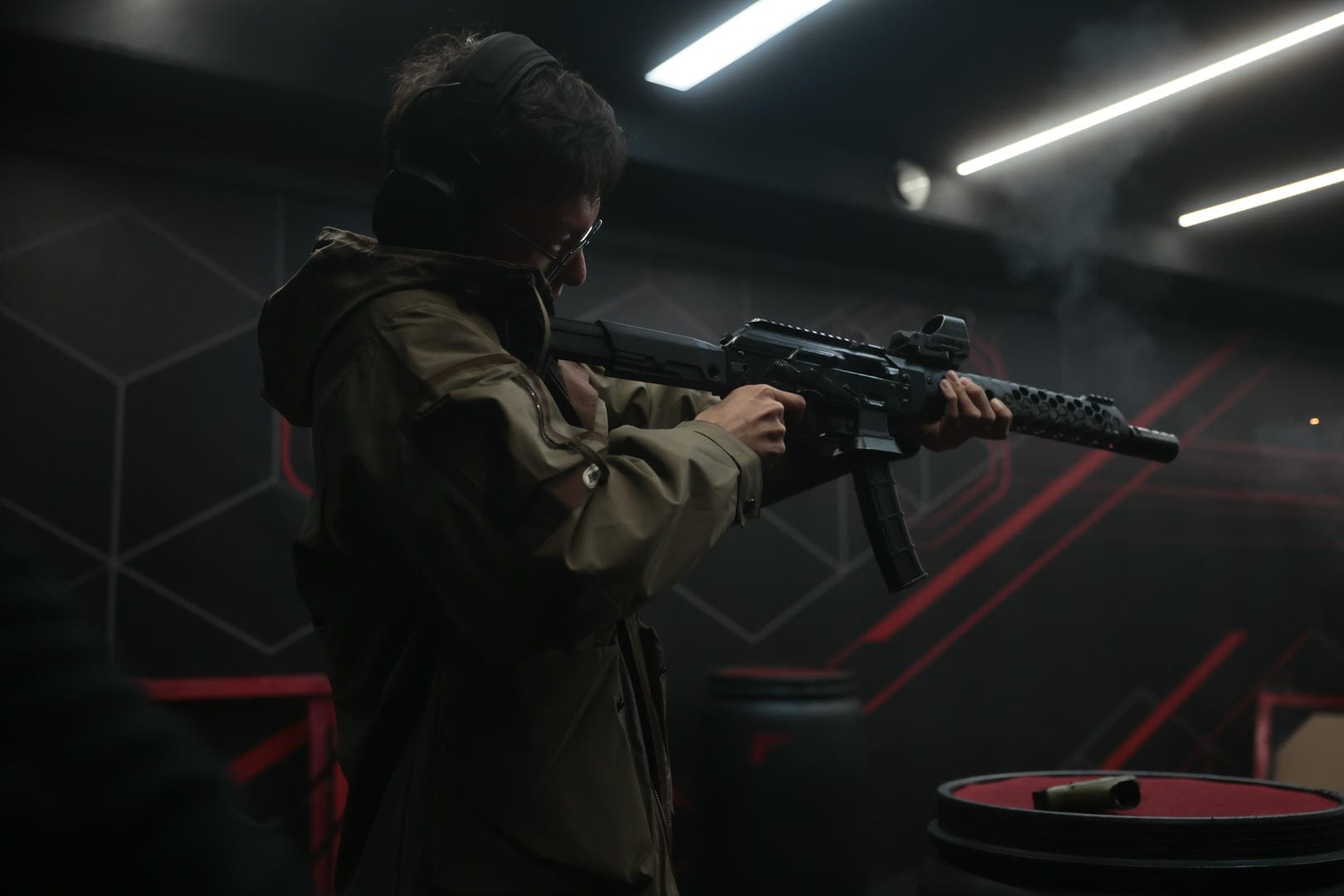 A person wearing headphones and glasses aiming a rifle at an indoor shooting range.