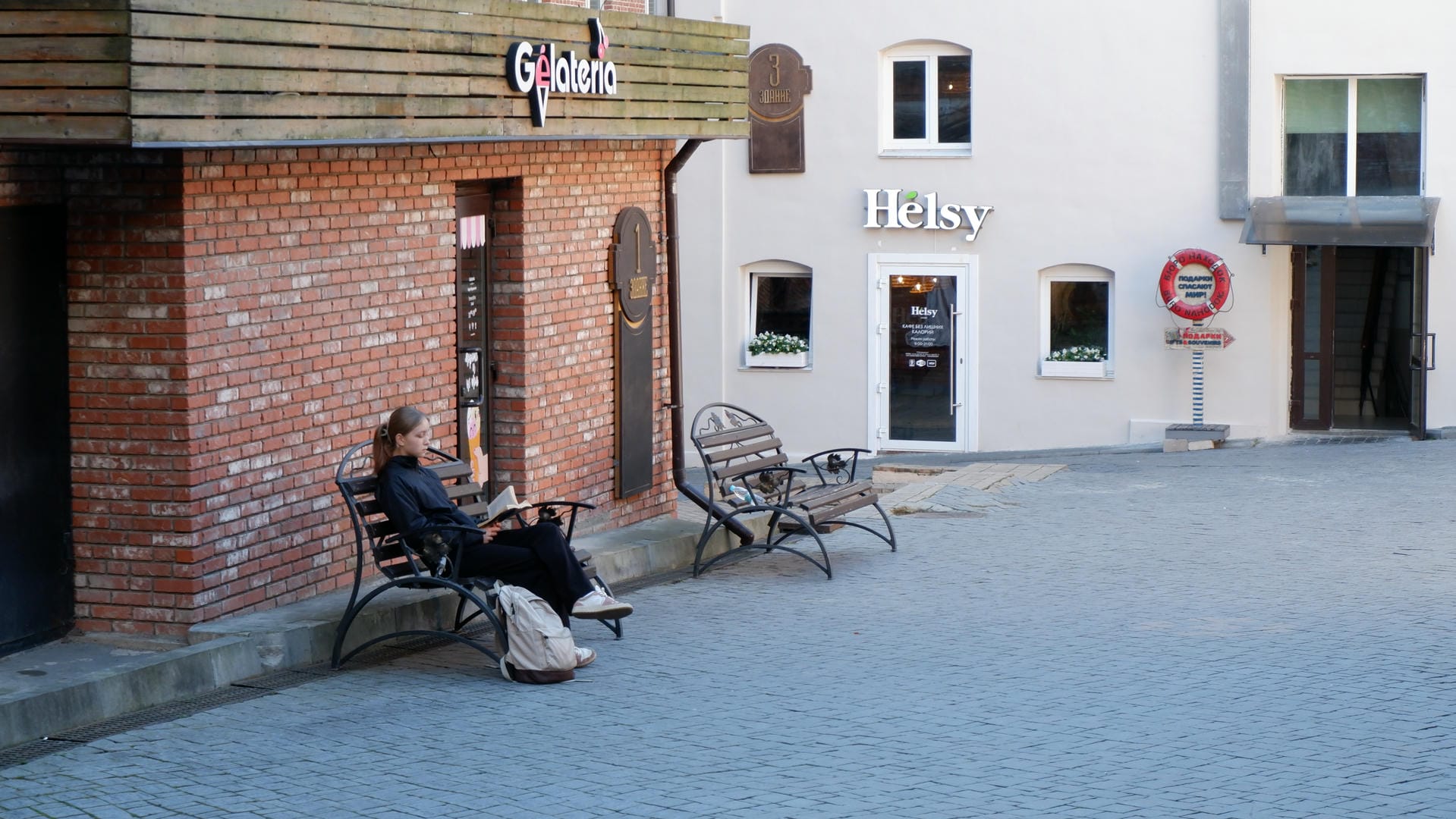 A person sitting on an outdoor bench with the storefronts of Gelateria and Helsy visible nearby.
