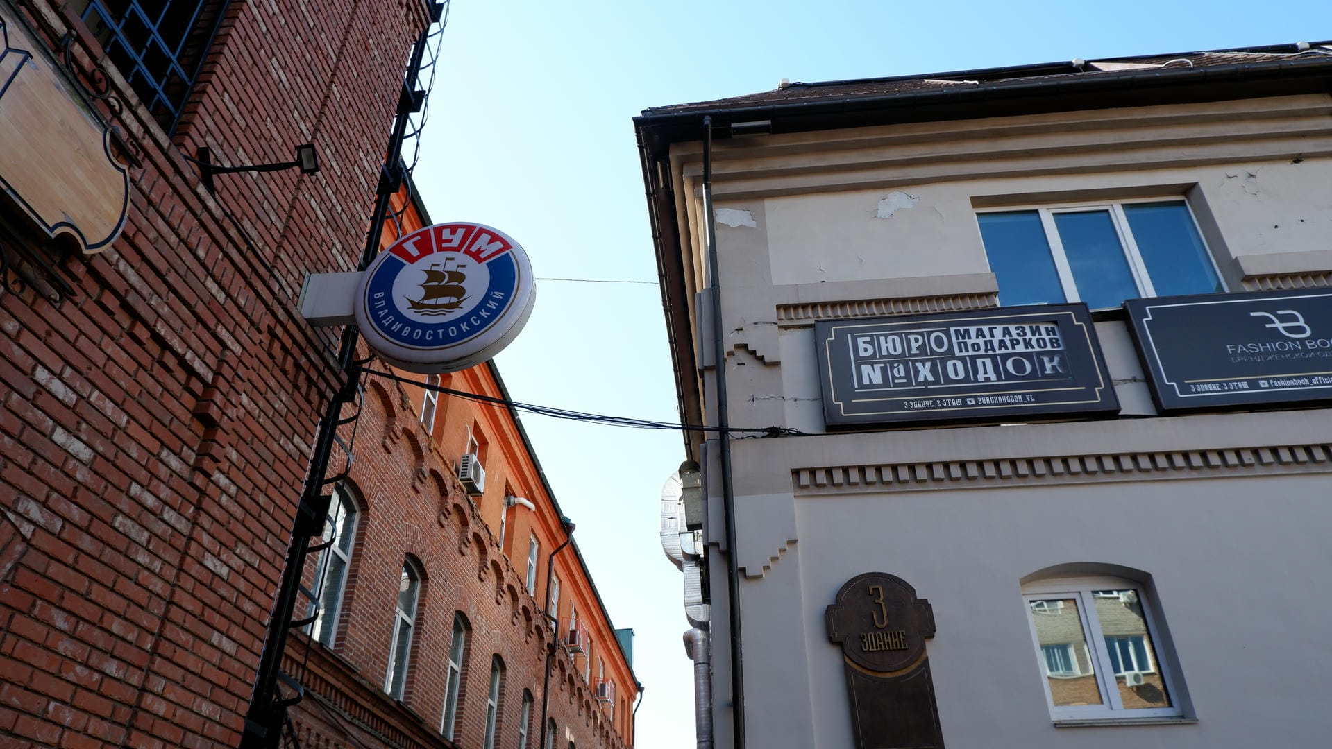 Buildings with Russian signs on the street, including “ＢＵＲＯ STORE ＧＩＦＴＳ”.