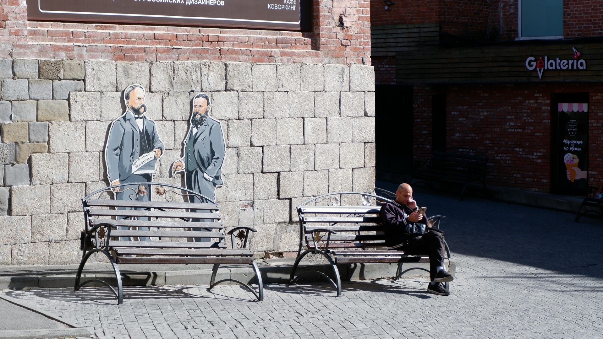 A person sitting on a bench using a phone, with a mural of two suited figures on the wall behind them.