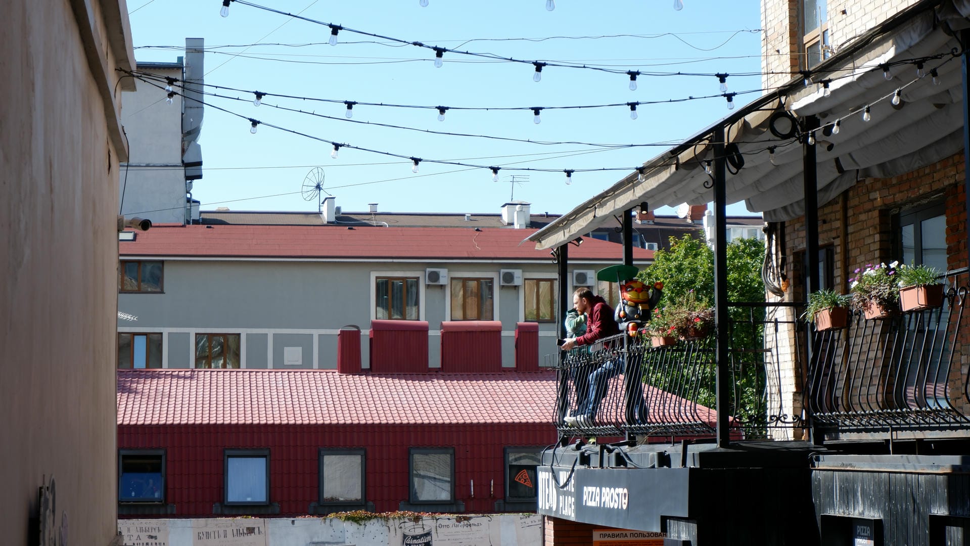 Two people standing on a balcony surrounded by potted plants and string lights.